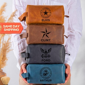 May include: Four personalized leather toiletry bags stacked on top of each other. The top bag is brown with a U.S. Army veteran logo and the name "Elvis". The second bag is brown with a star and the name "Clint". The third bag is dark gray with a military eagle and the name "Ford". The bottom bag is blue with a U.S. Marine Corps veteran logo and the name "Arthur".