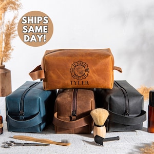 May include: A collection of leather toiletry bags in various colors: brown, blue, and black. The top bag is embossed with a fire department emblem and the name "Tyler." Each bag has a zipper closure and a handle. A shaving brush, scissors, and toothbrush are in the foreground.