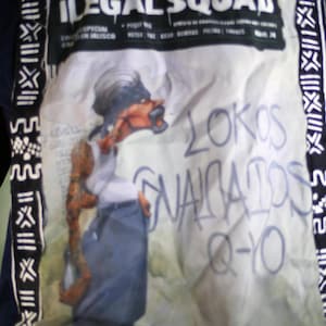 May include: A white t-shirt with a black and white graphic print of a cartoon character. The text "Illegal Squad" is printed in black at the top of the graphic. The text "Lokos Enajenados Q-Yo" is printed in black at the bottom of the graphic.