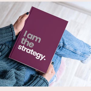 Puede incluir: Un cuaderno burdeos con el texto "I am the strategy" en letras plateadas. El cuaderno lo sostiene una persona que lleva un suéter verde azulado y vaqueros azules.