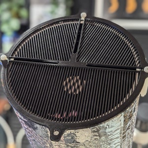 Op de afbeelding: Een zwarte metalen grill met drie secties, elk met een aparte handgreep. De grill is ontworpen om over een ronde pot of pan te passen.