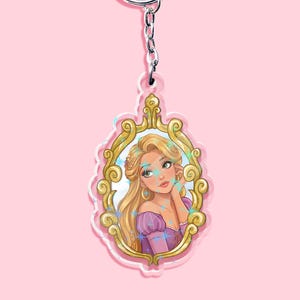 May include: A Rapunzel-themed keychain featuring a portrait of the character within a gold-colored ornate frame. The keychain has a silver-colored ring and chain. The background is a solid light pink.