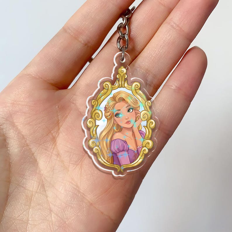 May include: A clear acrylic keychain featuring a cartoon illustration of a princess with long blonde hair, wearing a purple dress, and framed by a gold ornate border. The keychain has a silver chain.