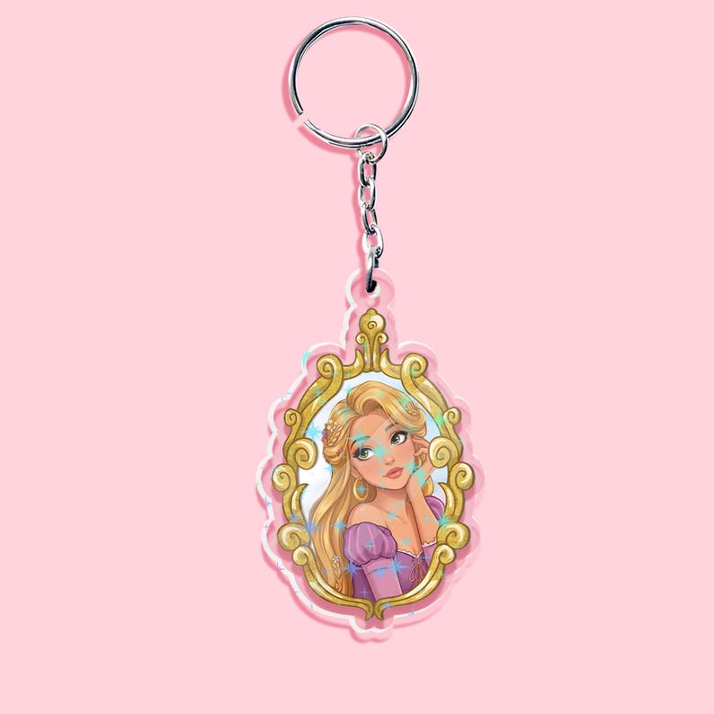May include: A Rapunzel-themed keychain featuring a portrait of the character within a gold-colored ornate frame. The keychain has a silver-colored ring and chain. The background is a solid light pink.