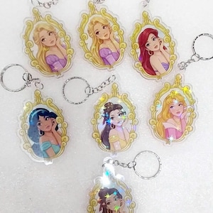 May include: Keychains featuring various cartoon princesses in ornate gold frames. Each keychain has a silver keyring attached. The princesses have different hair colors and wear different colored dresses. The keychains are made of clear plastic.