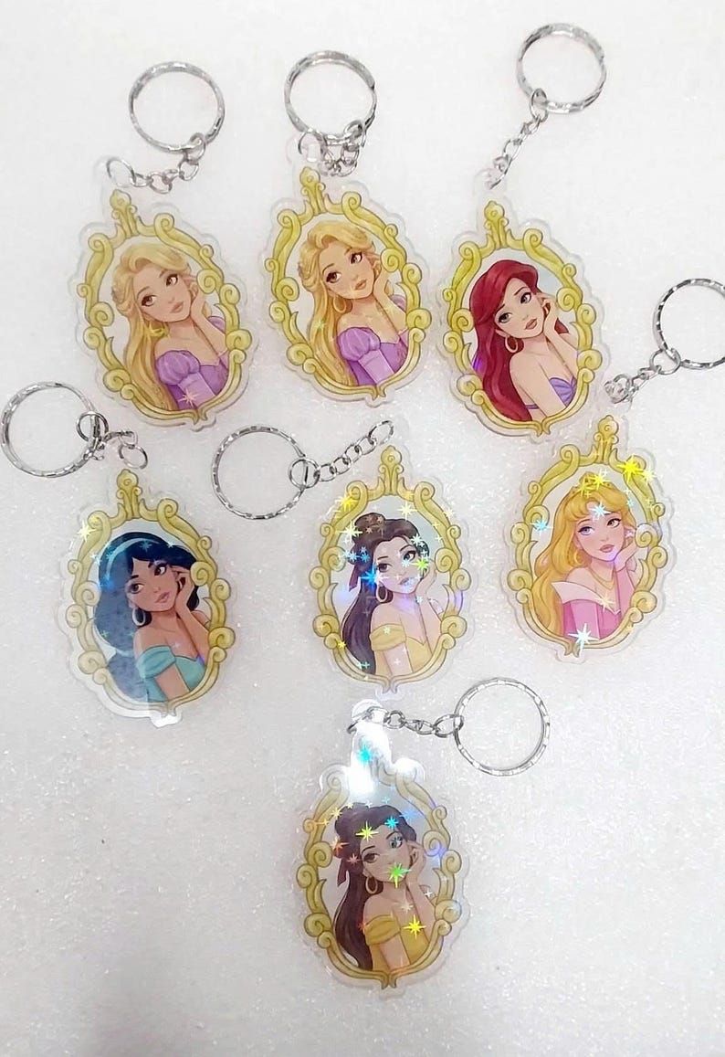 May include: Keychains featuring various cartoon princesses in ornate gold frames. Each keychain has a silver keyring attached. The princesses have different hair colors and wear different colored dresses. The keychains are made of clear plastic.