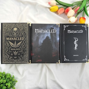 May include: Three hardback books with the title "Manacled" in a gothic font. The covers feature intricate designs, including a skull, a hooded figure, and a snake with a sword. Gold corner accents add a touch of elegance.