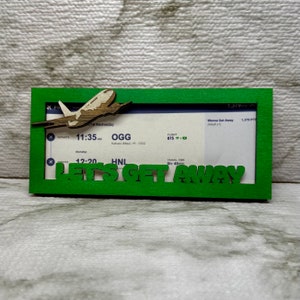 May include: A green wooden frame with a cutout of a plane and the text "Let's Get Away". The frame includes a mock airline ticket with the text "Wanna Get Away" and flight details for a flight from OGG to HNL.