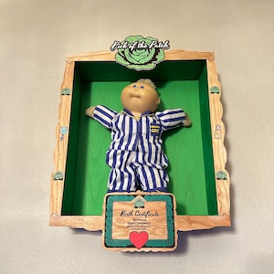 Könnte beinhalten: Eine Cabbage Patch Kids-Puppe in blau-weiß gestreiftem Pyjama, ausgestellt in einer Holz-Schattenbox mit grünem Innenraum. Die Box enthält eine Geburtsurkunde und ein "Pick of the Patch"-Logo.