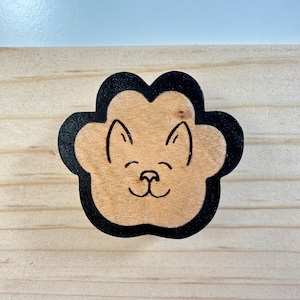 May include: A wooden paw print-shaped decoration with a smiling cat face etched into the center. The cat face is light brown, with black outlines and details. The paw print has a black border and is set against a light wood background.