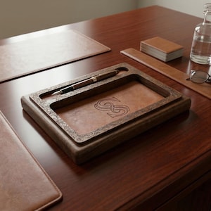 May include: A wooden desk organizer with a brown leather insert, holding two pens. The organizer has a decorative floral border and an engraved monogram. Other desk accessories include a leather blotter, a ruler, and a stack of cards.