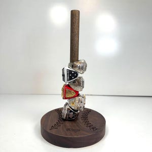 May include: A wooden ring holder with a dark brown base and a vertical wooden dowel. Five championship rings are stacked on the dowel. The rings are silver and gold with various colored accents and text, including the word "FINALIST".