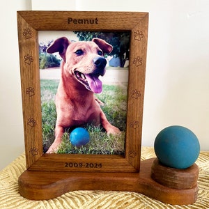 Könnte beinhalten: Ein hölzerner Gedenkrahmen für Haustiere mit einem Foto eines braunen Hundes und dem Namen "Peanut" eingraviert oben. Der Rahmen hat Pfotenabdrücke an den Seiten und die Daten "2009-2024" unten eingraviert. Ein blauer Ball befindet sich auf einem Holzsockel neben dem Rahmen.