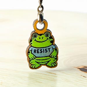 May include: A wooden frog charm with a light blue shirt that says "RESIST". The frog is green with black spots and a smiling face. The charm is attached to a silver-colored metal loop.