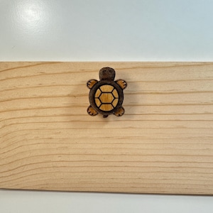 May include: A wooden turtle-shaped decorative item with a dark brown body and a lighter brown shell. The turtle is centered on a light-colored wooden surface with visible wood grain. The background is white.