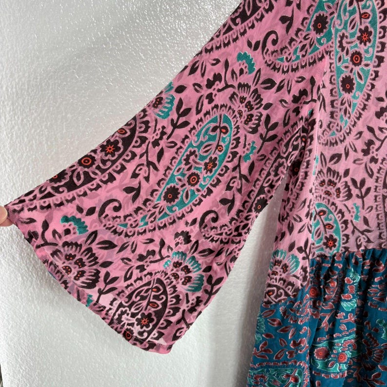 Op de afbeelding: Een roze en teal paisley print jurk met klokmouwen. De jurk heeft een gedetailleerd bloemen- en paisley patroon in roze, teal en bruine tinten. De onderkant van de jurk is teal met een vergelijkbaar paisley design.