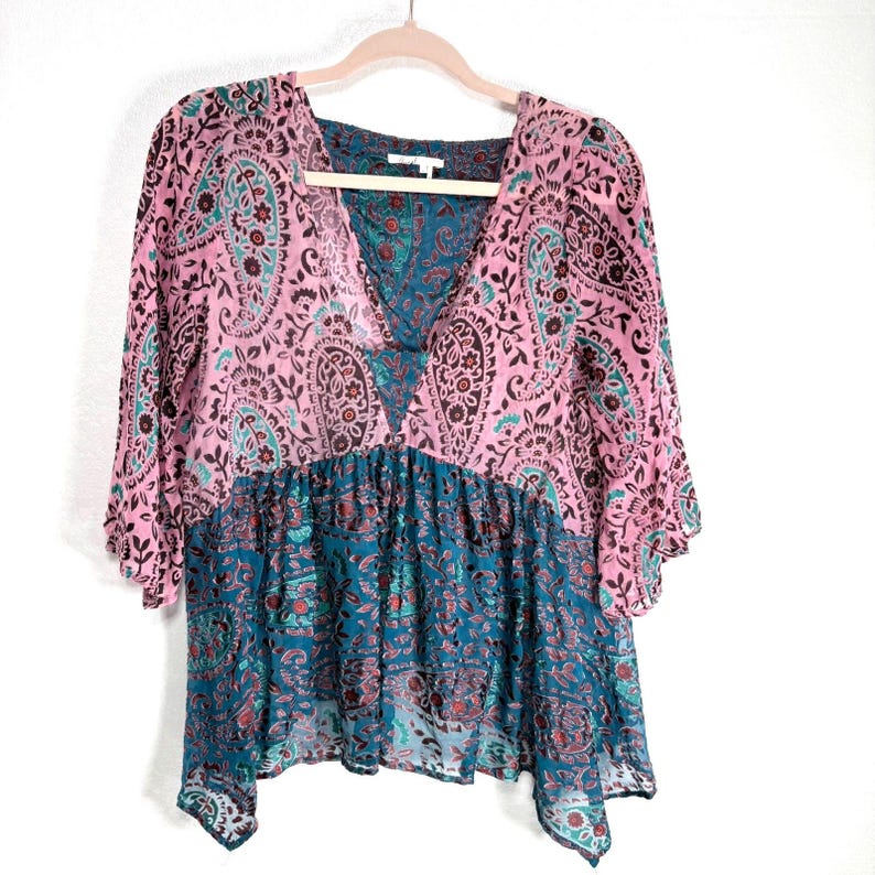 Op de afbeelding: Een roze en teal paisley print top met een V-hals en korte mouwen. De top heeft een vloeiend, gelaagd ontwerp met een mix van bloemen- en paisley patronen. De top hangt aan een houten hanger.