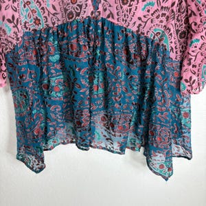 Op de afbeelding: Een roze en teal top met een patroon en een V-hals. Het ontwerp bevat bloemen- en paisley-motieven. De stof lijkt licht en vloeiend, met een gelaagde constructie. Het kleurenpalet omvat roze, teal en bordeaux.