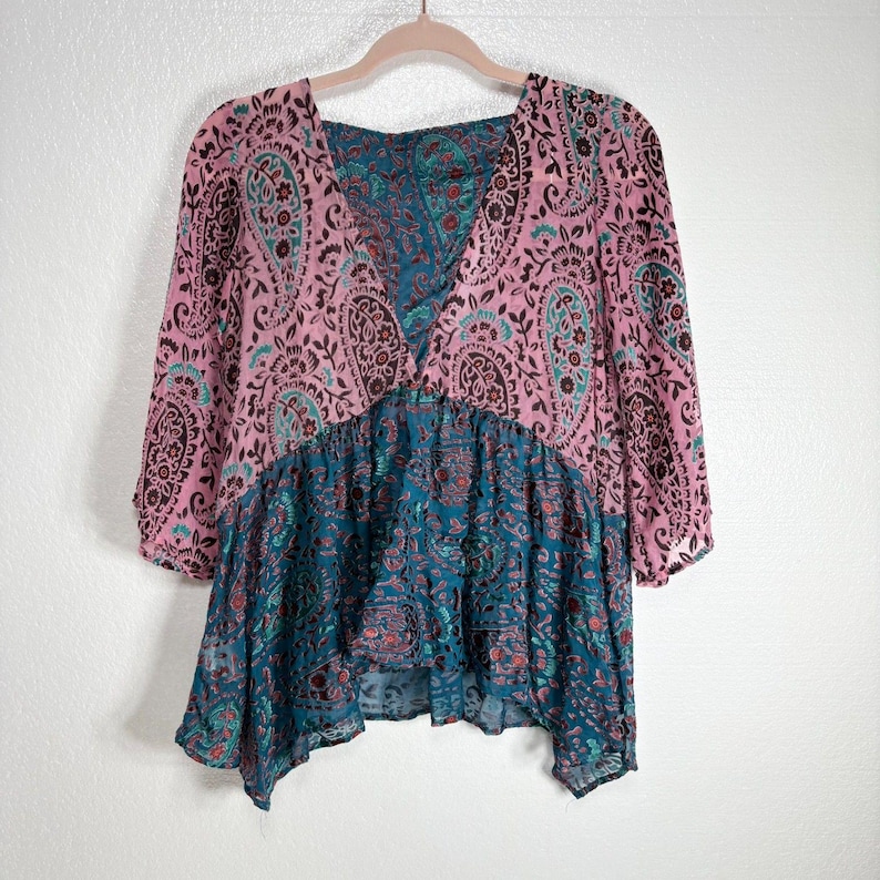 Op de afbeelding: Een roze en teal paisley print top met een V-hals en driekwart mouwen. De top heeft een vloeiende, asymmetrische zoom. Het roze gedeelte heeft een paisley patroon met bruine en teal accenten, terwijl het onderste gedeelte teal is met een vergelijkbaar ontwerp.
