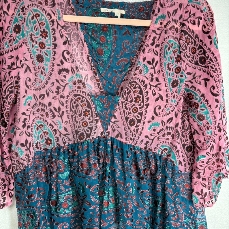 Op de afbeelding: Een V-hals jurk met een paisley patroon in roze en teal. De jurk heeft driekwart mouwen en een gerimpelde taille. De stof lijkt licht en vloeiend, geschikt voor warm weer.