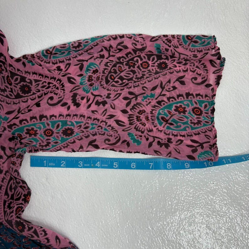 Op de afbeelding: Close-up van een roze paisley-stof met teal en bruine accenten. Een blauwe meetlint is langs de rand van de stof geplaatst, metingen in centimeters.