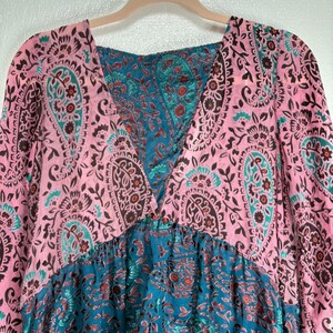 Op de afbeelding: Een roze en teal paisley print jurk met een V-hals en lange mouwen. De jurk heeft een combinatie van roze en teal paisley patronen, met een geplooide taille en een vloeiend silhouet.
