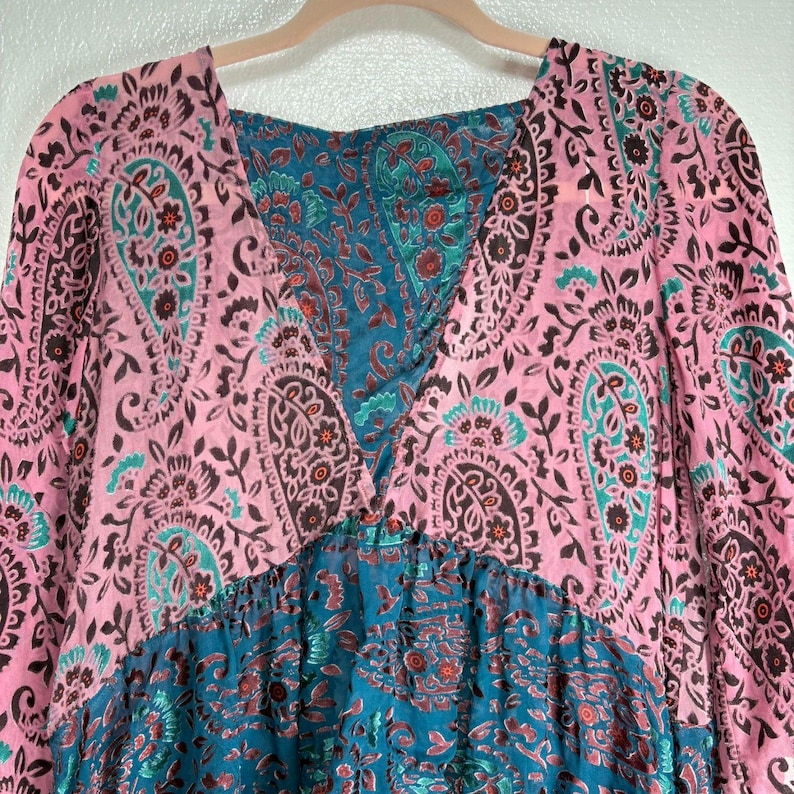 Op de afbeelding: Een roze en teal paisley print jurk met een V-hals en lange mouwen. De jurk heeft een combinatie van roze en teal paisley patronen, met een geplooide taille en een vloeiend silhouet.