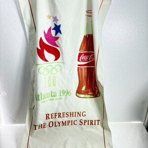 May include: A white rectangular kitchen towel featuring a red Coca-Cola bottle graphic, the 1996 Atlanta Olympic torch logo with five rings, and the text "Atlanta 1996" and "REFRESHING THE OLYMPIC SPIRIT" printed in red and green ink on the fabric.