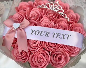 May include: A bouquet of pink roses, wrapped in decorative paper and ribbon. A silver tiara sits atop the roses, and a banner with the text "YOUR TEXT" is across the front. The bouquet is tied with a pink bow.