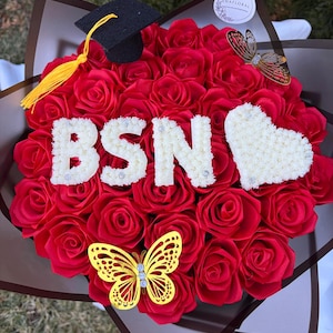 Peut inclure: Un bouquet de roses rouges avec les lettres "BSN" en blanc, orné d'une toque de graduation et d'une composition florale en forme de cœur. Des papillons dorés décorent l'ensemble. Un design floral de célébration.