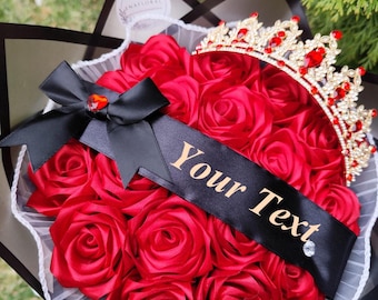 May include: A bouquet of red satin roses, topped with a gold and red jeweled crown. A black ribbon with the text "Your Text" is draped across the roses. The bouquet is wrapped in black and white paper.