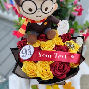 May include: A plush doll with round glasses and a lightning bolt scar sits atop a bouquet of artificial yellow and maroon roses. A red ribbon across the flowers features the text 9 3/4 and Your Text, accompanied by a spiderweb charm.
