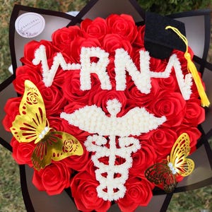 May include: A floral arrangement of red roses with white RN lettering and a Caduceus symbol. Includes a small graduation cap and gold butterfly accents. The arrangement is presented in a decorative box.