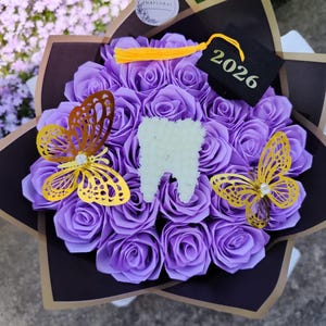 May include: A bouquet of purple roses with gold butterfly accents and a tooth-shaped centerpiece. A small graduation cap with the year 2026 is also included. The bouquet is wrapped in black and brown paper.