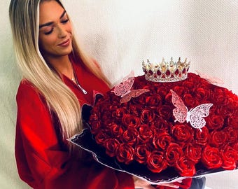 May include: A large bouquet of deep red roses wrapped in black paper, featuring a metallic silver crown with red accents resting on top and two white decorative butterfly cutouts attached to the floral arrangement with a red ribbon.