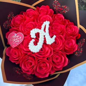 Può includere: Un bouquet di rose rosse con una "A" floreale bianca al centro. Il bouquet è avvolto in carta marrone scuro con finiture dorate e dettagli a forma di cuore con le parole "Love" e "Happy Valentine's Day". Il bouquet è di ENAFLORAL.
