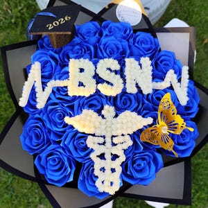 May include: A bouquet of blue roses with the letters "BSN" in white, a caduceus symbol, and a gold butterfly. A small graduation cap with "2026" is also included. The bouquet is wrapped in black and grey paper.