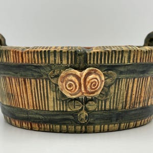 May include: A handcrafted ceramic planter with a barrel design. The planter features vertical lines in shades of tan and brown, accented with dark green bands. Two handles and a floral embellishment add detail to this decorative piece.