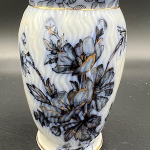 May include: A white ceramic vase with a flared top and a gold rim. The vase features a blue and gold floral design. The body of the vase has a textured, wavy pattern. The vase is a decorative item.