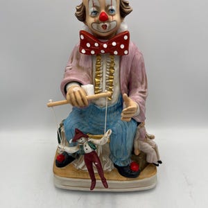 May include: A ceramic clown figurine wearing a red top hat and a bow tie with white spots. The clown is seated and holding a puppet. The puppet is dressed in red trousers and a green waistcoat. The clown's outfit is pink and blue.