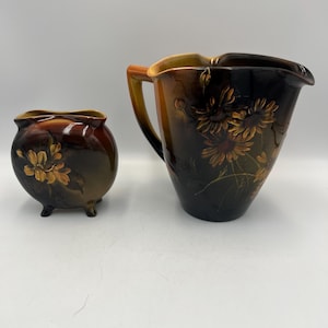 May include: A ceramic pitcher and small bowl set. The pitcher has a handle and a spout, with a floral design. The bowl has three small feet. Both pieces are glazed in shades of brown and gold.