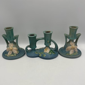May include: Three teal ceramic candle holders with floral and leaf designs. The candle holders have a matte finish and are in various shapes, including a double-holder and two single holders. The floral accents are in shades of cream and pink.