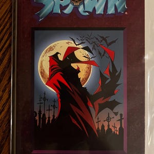 May include: A comic book cover featuring the word "SPAWN" in blue, with a skull icon. The artwork depicts a figure in a red cape against a full moon, with bats and a graveyard. The author's name, "TODD MCFARLANE," is at the bottom.