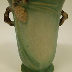 May include: A green ceramic vase with a flared top and a round base. The vase features two handles and a pinecone design on the side. The vase is approximately 10 inches tall.