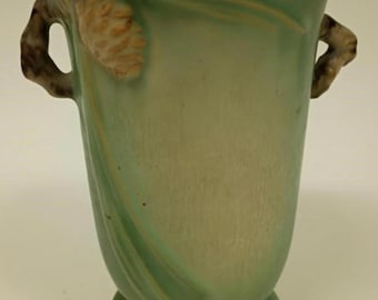 Może przedstawiać: Zielony ceramiczny wazon z rozkloszowaną górą i okrągłą podstawą. Wazon posiada dwa uchwyty i wzór szyszki sosnowej z boku. Wazon ma około 25 cm wysokości.