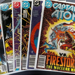 Puede incluir: Una colección de cómics vintage de DC Comics, con Captain Atom y Firestorm. Las portadas muestran ilustraciones dinámicas de superhéroes, con texto en negrita y el logotipo de DC Comics. Los cómics están en fundas protectoras.