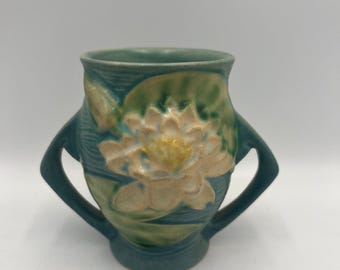 May include: A teal ceramic vase with two handles, featuring a raised design of a water lily. The flower is cream-colored with a yellow center, set against green lily pads. The vase has a textured surface and a slightly flared rim.