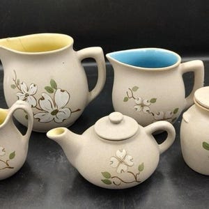 May include: A cream-colored ceramic tea set comprising a teapot, creamer, sugar bowl with lid, and two jugs. Each piece is adorned with a white floral pattern and green leaves. The teapot and jugs have handles.