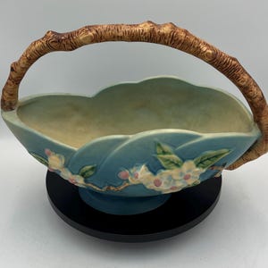 May include: A blue ceramic basket with a branch-like handle. The basket features a floral design with white and pink blossoms and green leaves. The handle is brown and textured. The basket is on a black base.