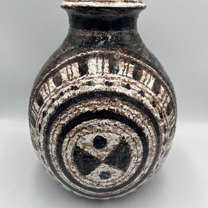 May include: A ceramic vase with a rounded body and a narrow neck. The vase features a textured surface with a pattern of concentric circles and geometric shapes in black, white, and brown. The vase is approximately 10 inches tall.